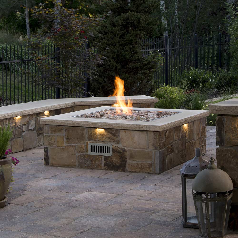 Square gas fire pit with flame burning in center over metal burner grate