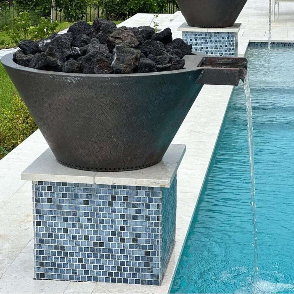 Round gas fire and water bowl in dark powder-coated metal with stone fill beside pool water.