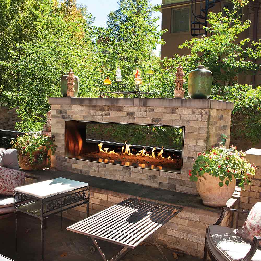 Outdoor linear fire pit fireplace with glass front and stone surround, flames visible