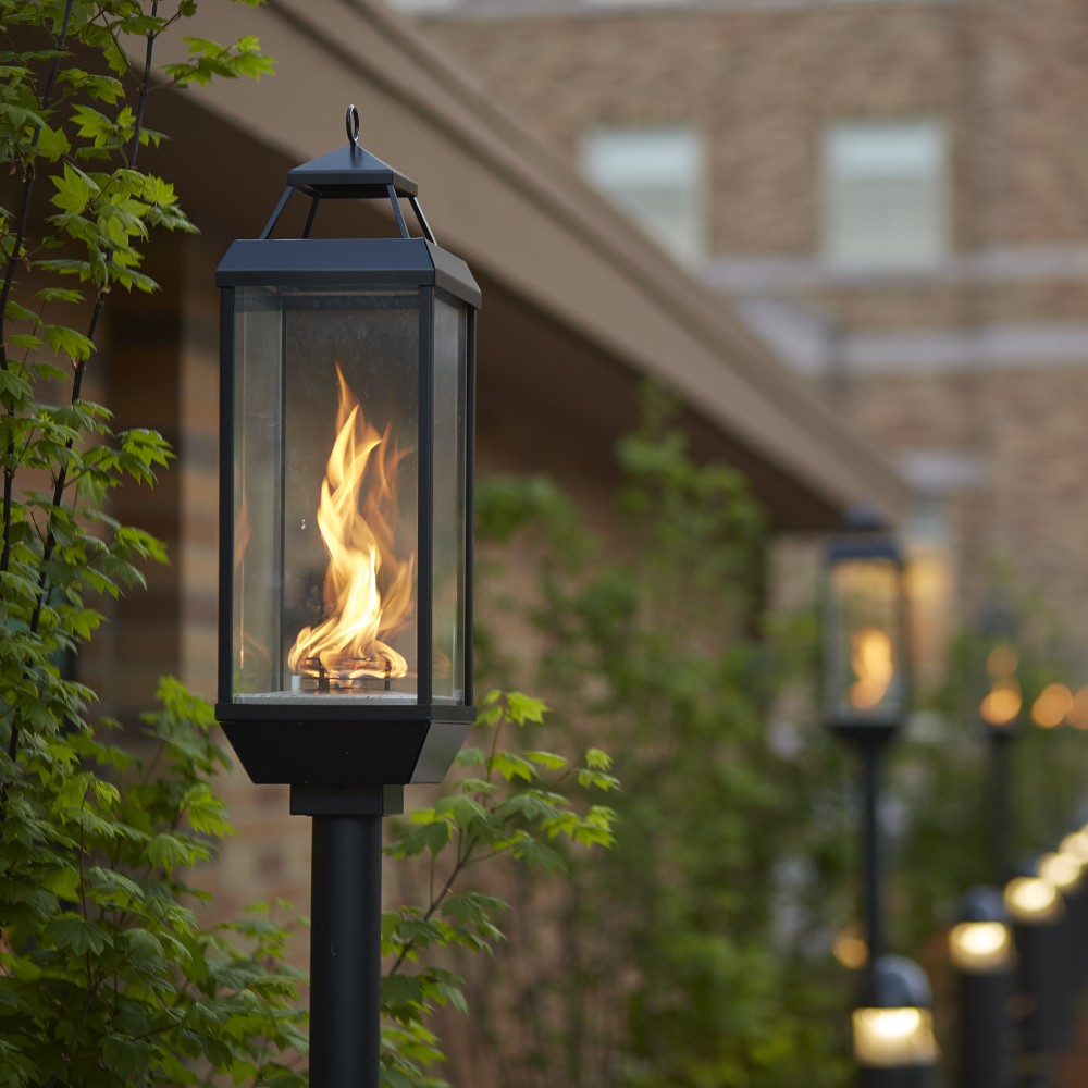 Black gas lantern on post with burning flame visible through clear glass and top loop