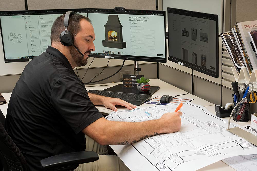Man working at a desk with multiple monitors and architectural drawings.